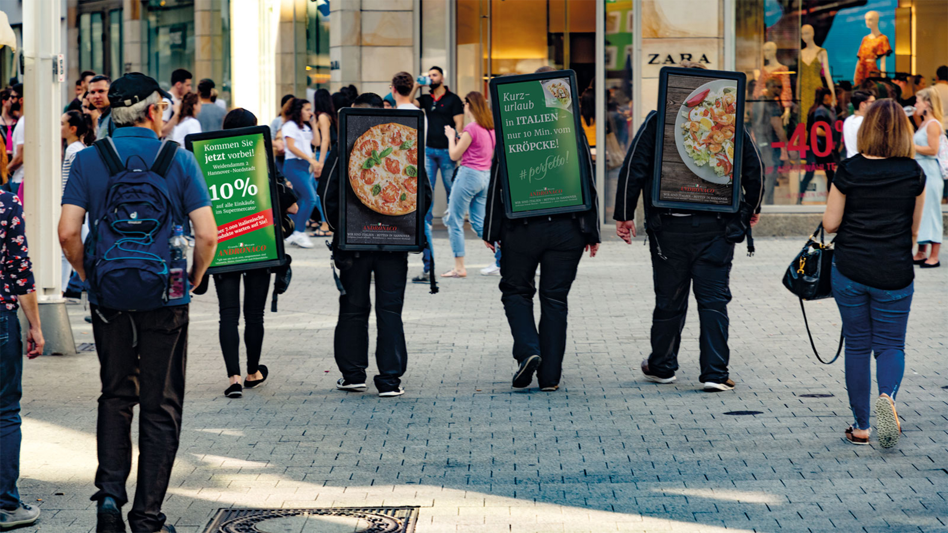 Mehrere Personen tragen DigiBoards von INOVISCO auf dem Rücken, die für Andronaco werben. Gezeigt werden Pizza, Pasta und Rabattaktionen in einer belebten Einkaufsstraße. Passanten flanieren direkt daneben durch die Fußgängerzone.
