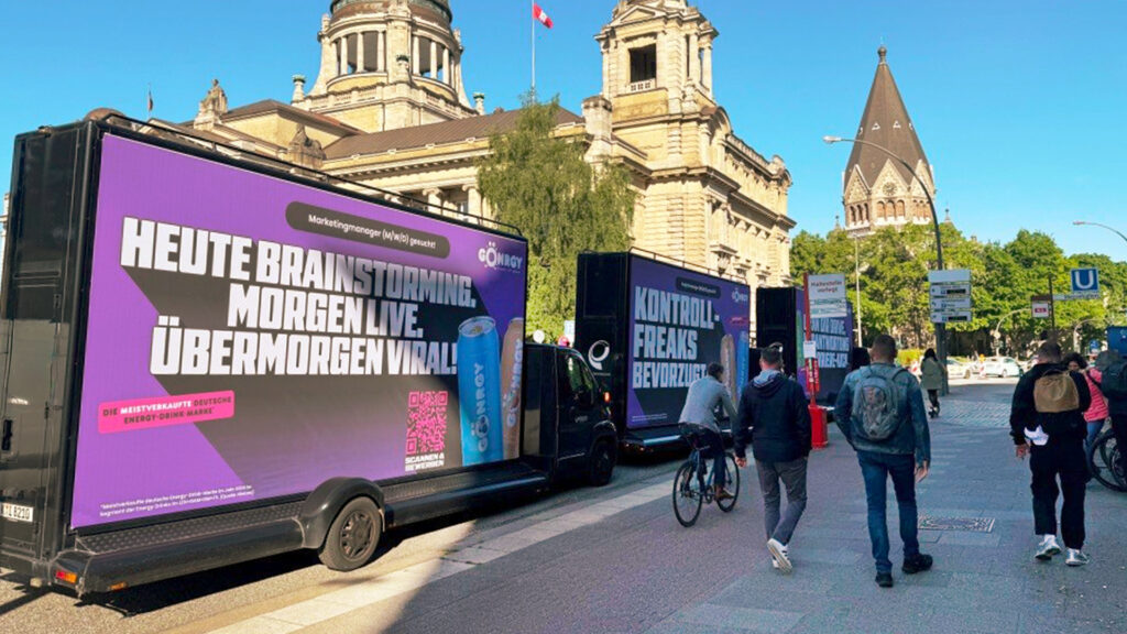 Auf dem Bild sind mehrere CoolLiteTrucks mit auffälliger Gönrgy-Werbung zu sehen. Links prangt der große Slogan: „HEUTE BRAINSTORMING. MORGEN LIVE. ÜBERMORGEN VIRAL!“ – daneben eine Getränkedose und ein QR-Code. Rechts daneben fährt ein weiterer Truck mit dem Claim: „KONTROLL-FREAKS BEVORZUGT“. Im Hintergrund erkennt man eine historische Architektur und ein Kirchengebäude, während Passanten und Radfahrer die Szene beleben.