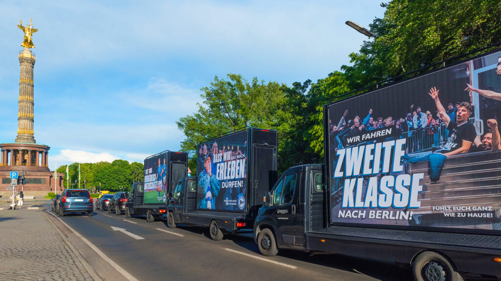Das Bild zeigt mehrere CoolLiteTrucks von INOVISCO, die in einer Reihe an der Berliner Siegessäule vorbeifahren. Auf den großen Werbeflächen der Trucks sind Fußball-Motive zu sehen: Vorne der Schriftzug „WIR FAHREN ZWEITE KLASSE NACH BERLIN!“ mit jubelnden Fans. Dahinter ein Truck mit der Botschaft „DASS WIR DAS NOCH ERLEBEN DÜRFEN!“. Weiter hinten ein Truck mit weiteren Fußball-Emotionen und Fans. Die Szenerie vermittelt ein starkes Fußball- und Fan-Feeling mitten in Berlin.