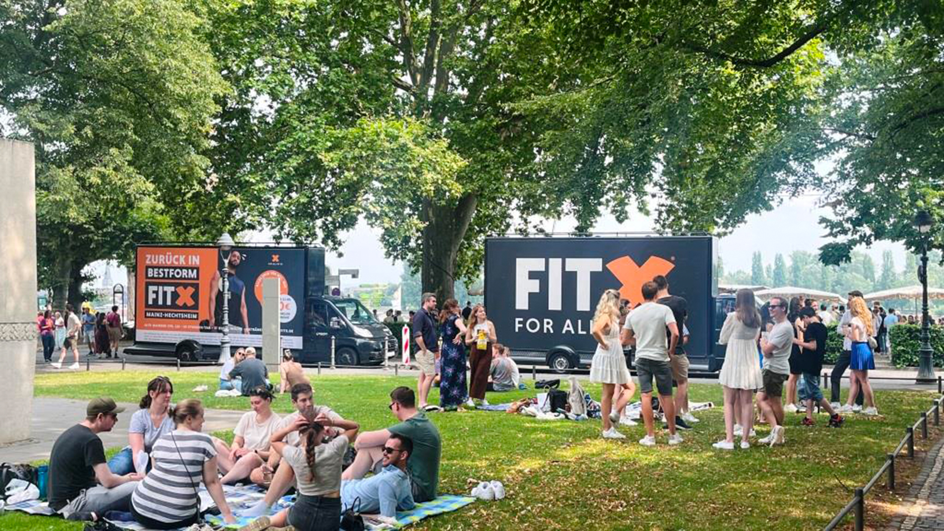 Zwei CoolLiteTrucks von INOVISCO stehen am Rand einer Grünfläche. Die großflächigen Werbemotive werben für das Fitnessstudio FitX mit Slogans wie „Zurück in Bestform“. Im Vordergrund sitzen und stehen viele junge Menschen bei sonnigem Wetter im Park.
