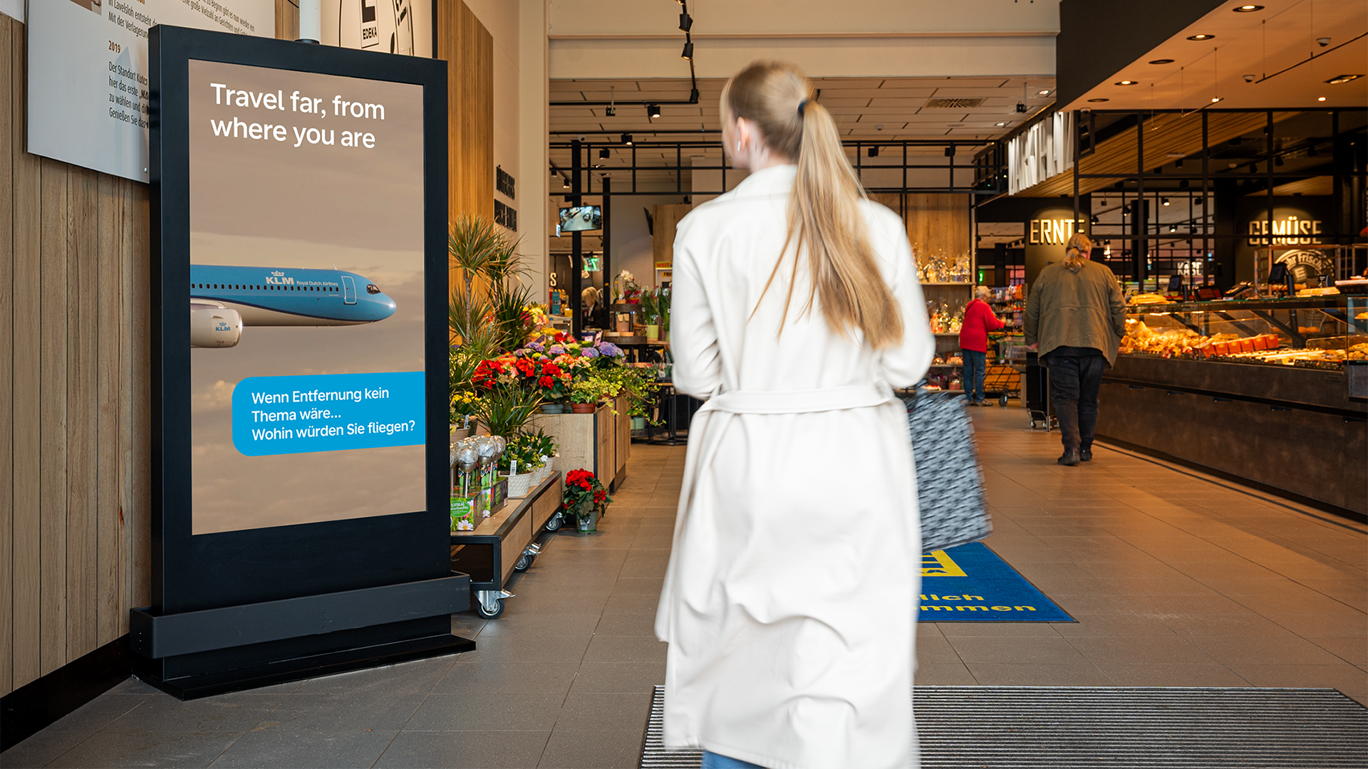 Ein digitales Citylight Poster (DCLP) im Eingangsbereich eines Supermarktes zeigt eine KLM-Werbung mit einem Flugzeug und dem Text „Travel far, from where you are“. Davor läuft eine Frau mit hellem Mantel an der Blumenabteilung vorbei in den Markt. Rechts sind frische Backwaren und Gemüseauslagen zu sehen.