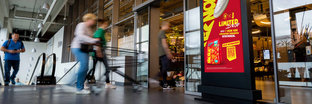 Ein INOVISCO DCLP steht am Eingang eines Supermarkts und zeigt eine auffällige Promotion für einen Sneaker-Gewinnspiel-Drop von NicNacs. Vorbeigehende Kundinnen und Kunden mit Einkaufswagen passieren die digitale Werbefläche.