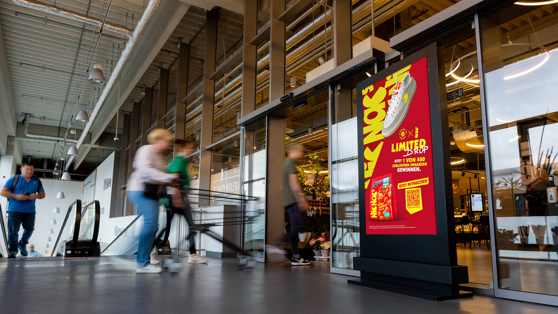 Ein INOVISCO DCLP im Eingangsbereich eines Supermarkts zeigt eine auffällige Promotion für einen Sneaker-Gewinnspiel-Drop. Kundinnen und Kunden mit Einkaufswagen passieren den digitalen Werbeträger auf dem Weg ins Geschäft.