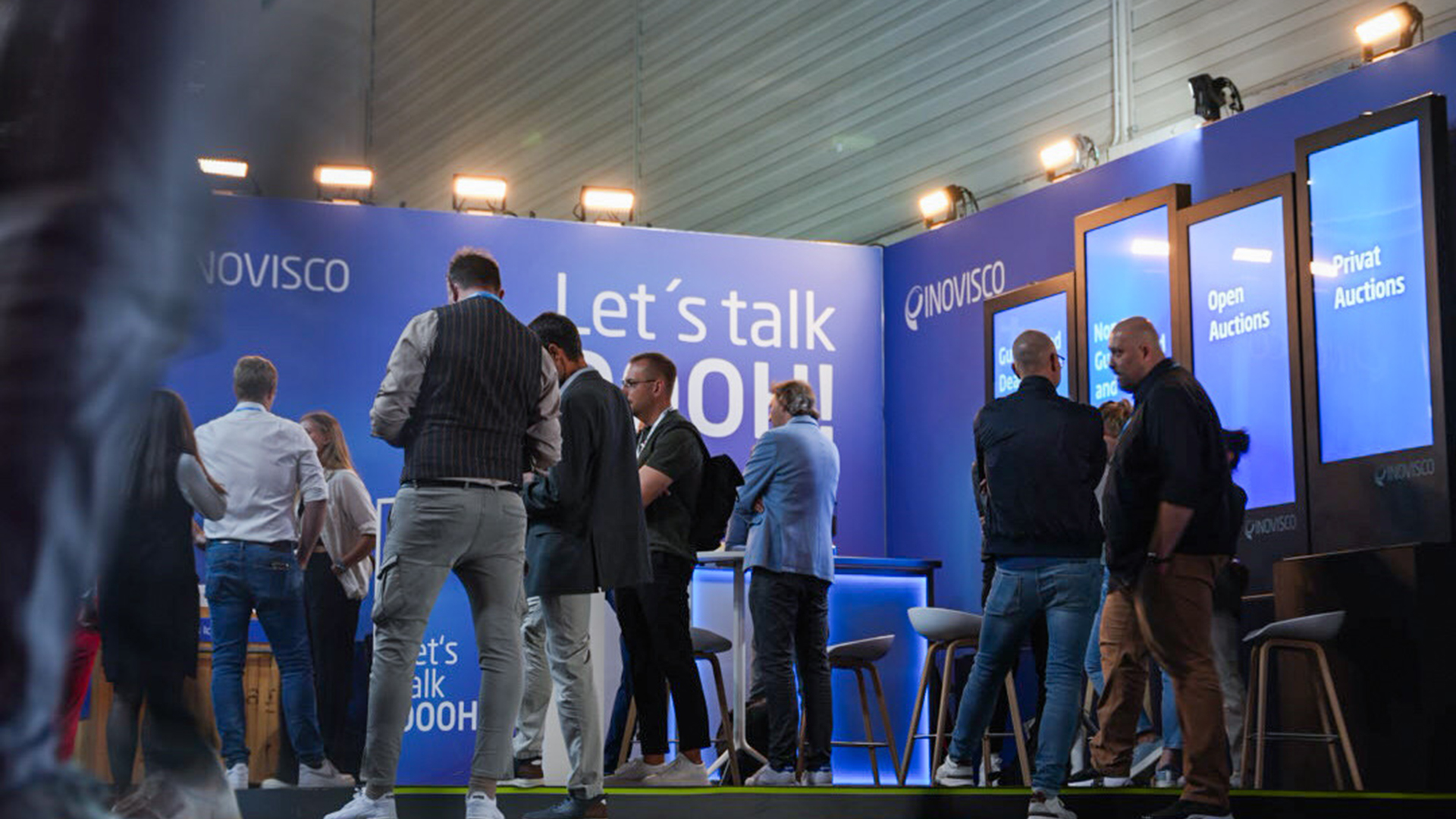 Besucherinnen und Besucher stehen und unterhalten sich am INOVISCO Messestand mit leuchtend blauer Wand und Screens. Der Schriftzug „Let’s talk DOOH!“ bildet den zentralen Blickfang.