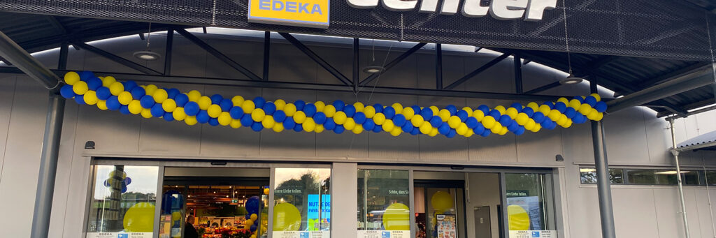 Eingangsbereich eines EDEKA-Marktes, dekoriert mit einer langen Kette aus blauen und gelben Luftballons. Durch die Glasfront sind Obst- und Gemüseabteilung sowie weitere Ballons erkennbar.