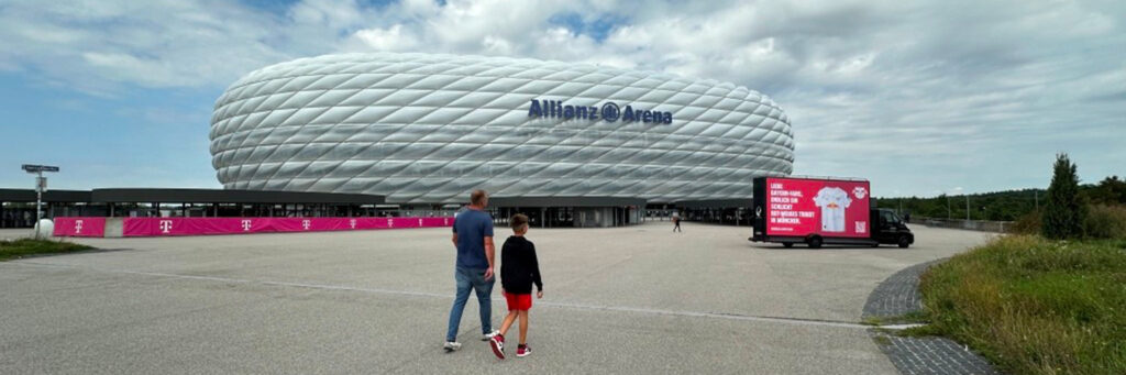 Vor der Allianz Arena steht ein INOVISCO CoolLiteTruck mit großflächiger, pinkfarbener Werbung. Zwei Personen gehen in Richtung Stadion, wodurch die mobile Werbefläche gut wahrgenommen wird. Das Medium ist direkt am Eingangsbereich platziert und dadurch besonders sichtbar.