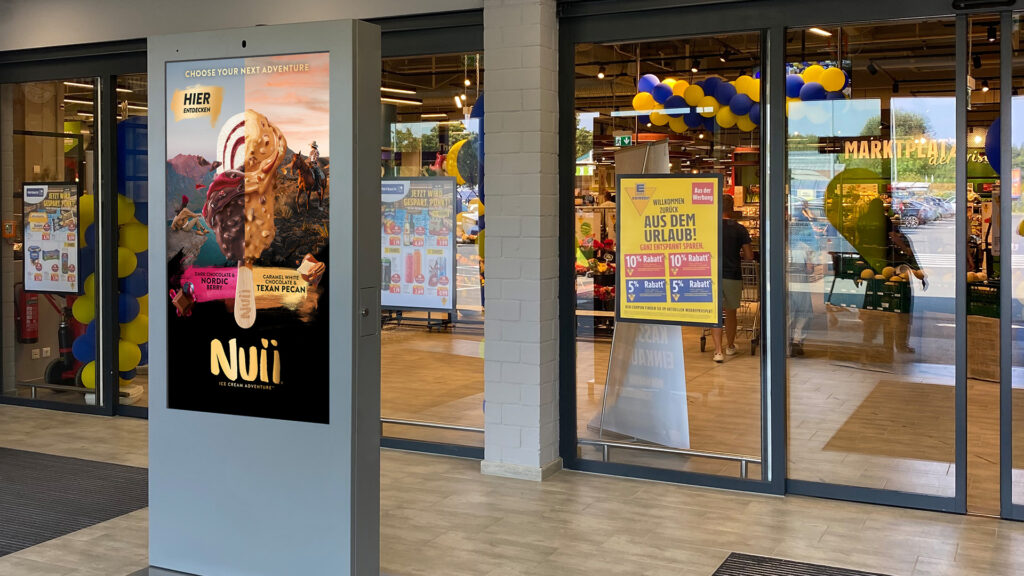 Ein digitales INOVISCO Display im Eingangsbereich eines EDEKA-Marktes zeigt eine Nuii-Eiswerbung. Im Hintergrund sind Einkaufswagen, Plakate und eine Ballondekoration in Blau-Gelb erkennbar.