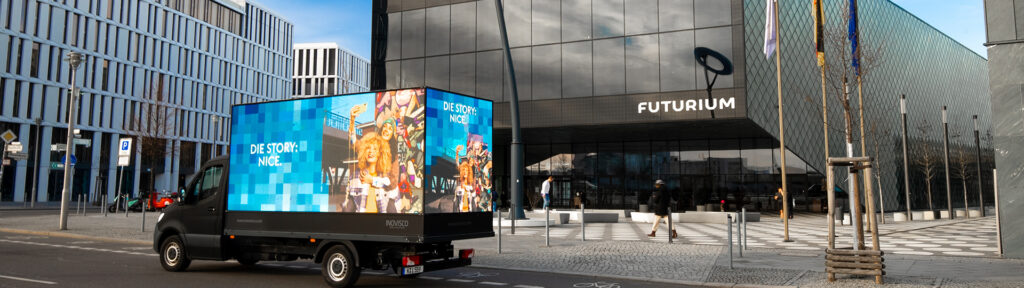 Ein DigiTruck von INOVISCO steht vor dem Futurium in Berlin. Auf den großen LED-Flächen ist eine bunte Werbung mit dem Schriftzug „Die Story: Nice.“ und fröhlichen Menschen zu sehen. Das moderne Glasgebäude spiegelt die Umgebung wider, während Passanten vorbeigehen.