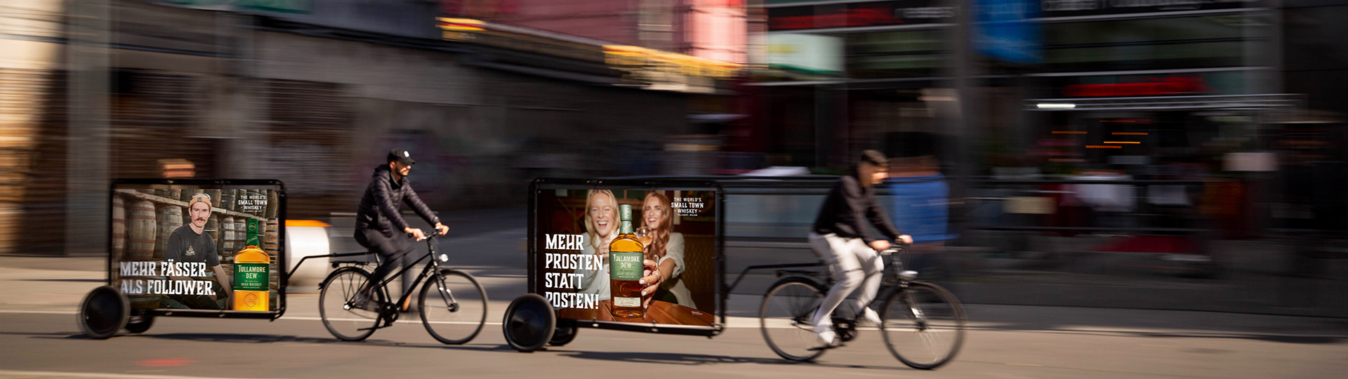 Auf dem Bild bewegen sich zwei Personen auf den INOVISCO-CoolLiteBikes nach rechts und ziehen die charakteristischen Werbetafeln hinter sich her. Auf den Tafeln ist eine Anzeige für Tullamore Dew Whiskey zu sehen.