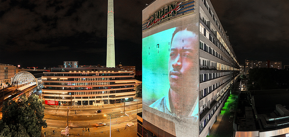 Aus einer Weitwinkel-Perspektive schaut man auf eine Straße, viele Gebäude und Skyline von Berlin. rechts wird eine Hausfassade von einem Digitalen Riesenposter von INOVISCO erleuchtet. Es wird eine Werbung der Marke "Tommy Hilfiger" an die Wand projiziert.