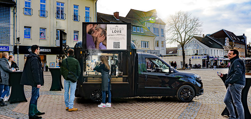 In einer Fußgängerzone steht ein INOVISCO DigiCoffeeVan, der eine Anzeige von PURELEI auf seinen Screens zeigt. Davor stehen einige Menschen, die einen Kaffee trinken und sich unterhalten.