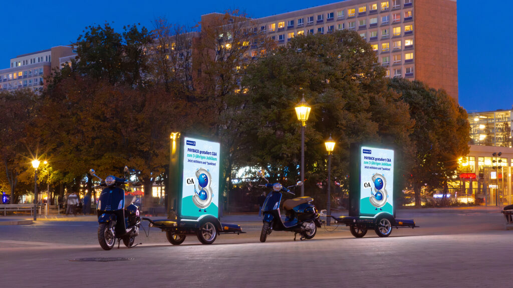 Zwei INOVISCO CoolLiteScooter stehen in der Abenddämmerung auf einem großen gepflasterten Platz und zeigen eine Werbung von Payback.