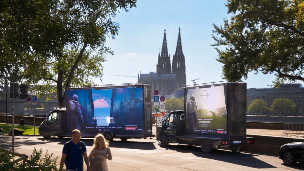 Auf dem Bild stehen zwei INOVISCO CoolLiteTrucks 36-1, die eine Werbung von dem Spiel "Starfield" zeigen. Im Hintergrund ist der Kölner Dom und blauer Himmel, im Vordergrund laufen zwei Personen vor den Trucks entlang.