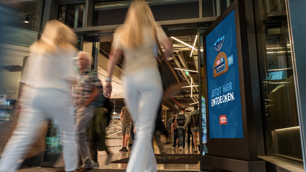 Zwei Personen laufen im Eingangsbereich eines Supermarktes an einem schwarzen DCLP von INOVISCO vorbei. Darauf ist eine Anzeige von Lieken Urkorn geschaltet.
