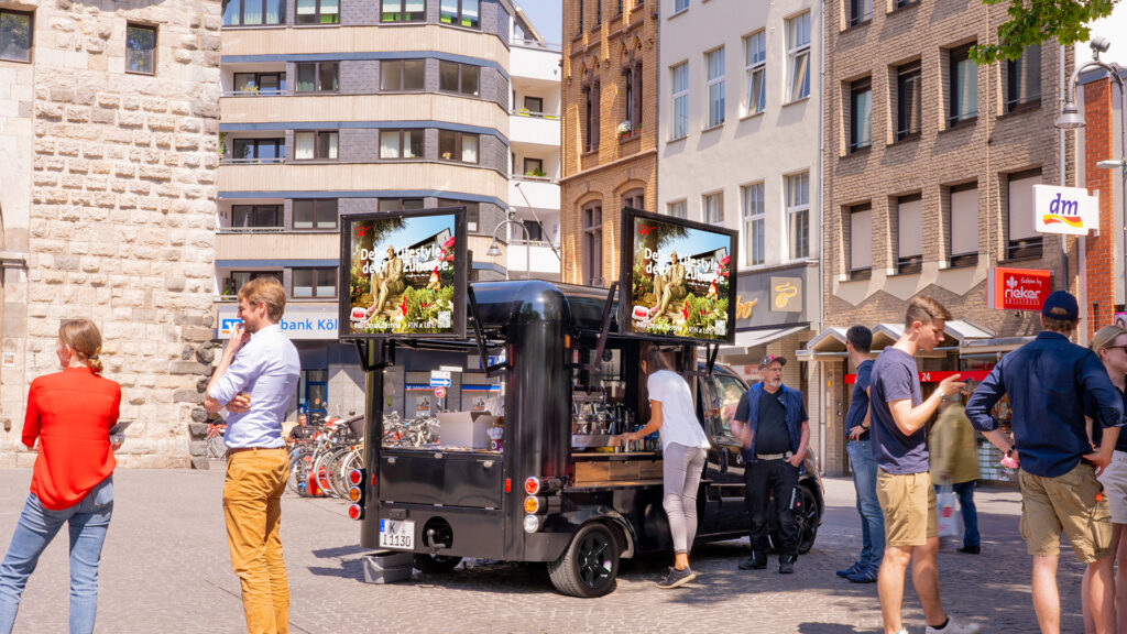 Eine Menge Menschen stehen um einen INOVISCO DigiCoffeeVan und unterhalten sich, während sie Kaffee trinken. Auf den Displays des Vans läuft Werbung von "LBS".