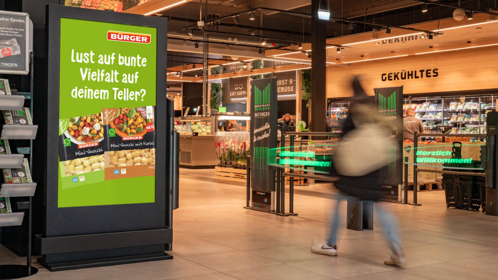 Links von einer Eingangsschranke steht ein schwarzer DCLP von INOVISCO. In den Markt geht gerade eine Person. Auf dem DCLP läuft eine Werbung von der Marke "Bürger".