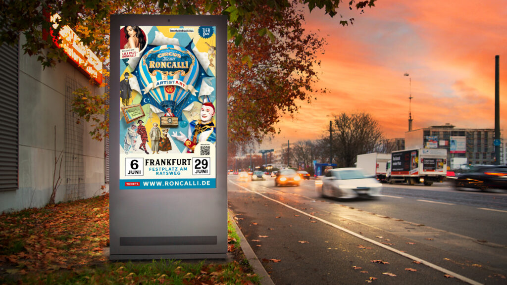 Ein digitales City-Light-Poster am Straßenrand zeigt eine bunte Zirkuswerbung von Roncalli. Im Hintergrund fahren Autos bei Abendstimmung vorbei.