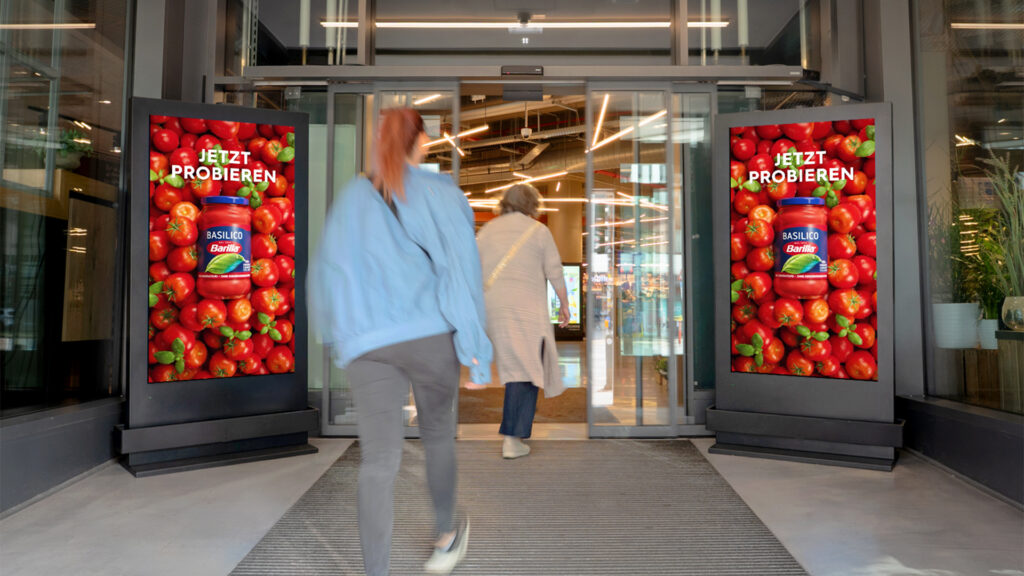 Zwei Frauen laufen hintereinander in einen Supermarkt. Links und rechts steht ein schwarzes DCLP mit einem Werbemotiv von Barilla.