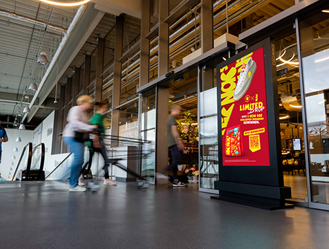 Drei Personen betreten einen Supermarkt. Rechts vom Eingang steht ein schwarzes DCLP mit einer NicNacs Werbung.