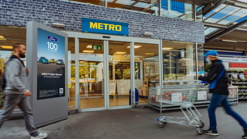 Zwei männliche Personen laufen in einen Metro Markt. Links steht ein graues INOVISCO DCLP mit einem Werbemotiv von Ford.