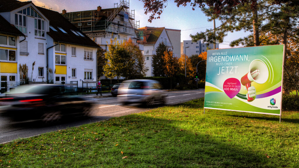 Ein Temporäres Großflächenplakat von INOVISCO steht am Straßenrand vor Wohnhäusern. Es zeigt eine bunte Unitymedia-Werbung mit Megafonmotiv und dem Slogan für Highspeed-Internet.