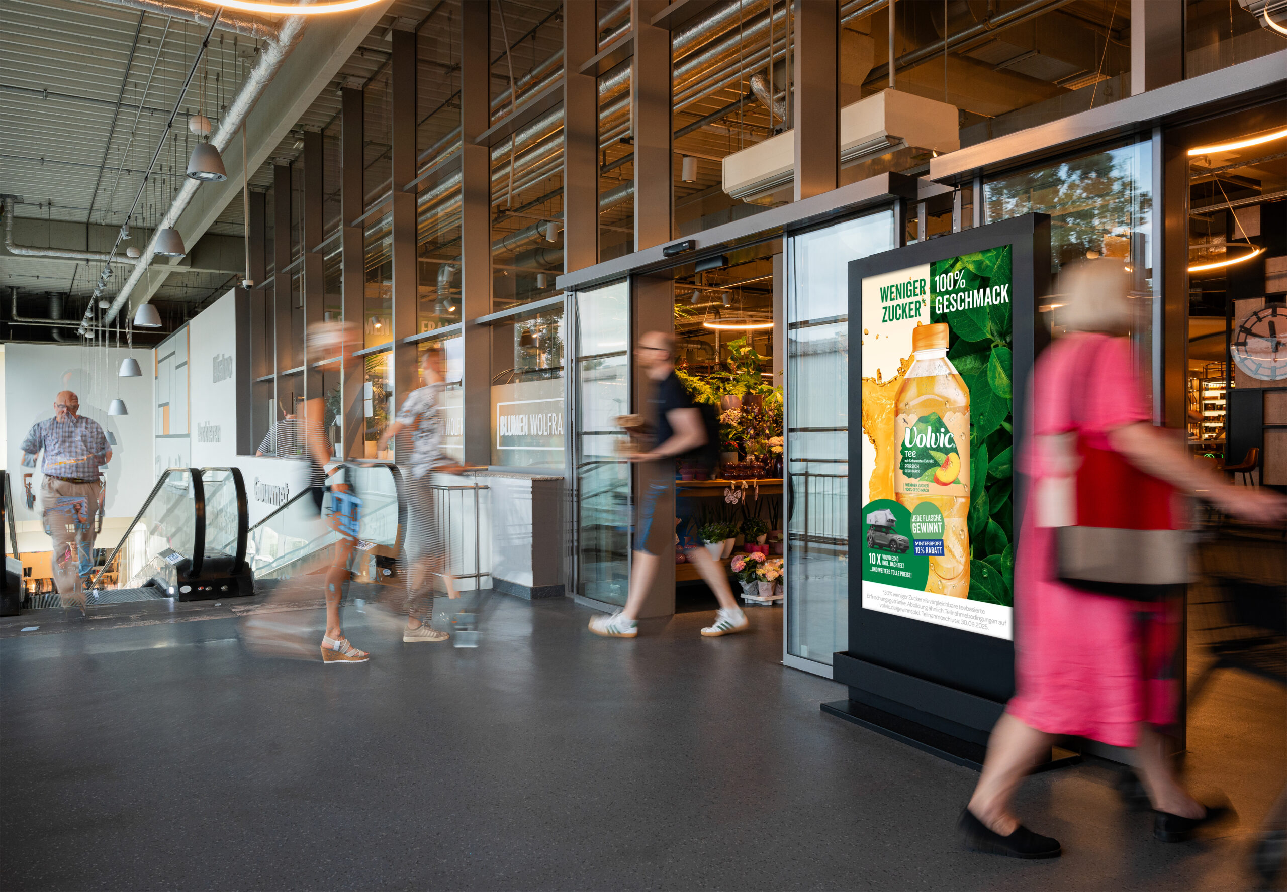 Ein digitales Citylight-Poster (DCLP) von INOVISCO im Eingangsbereich eines Supermarkts zeigt eine Volvic-Tee-Werbung. Mehrere Menschen gehen vorbei, während im Hintergrund Rolltreppen und ein Blumenladen erkennbar sind. Moderne, belebte Einkaufsatmosphäre.