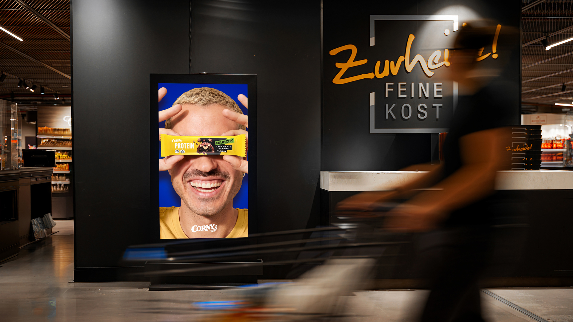Eine Person läuft mit Einkaufswagen an einem DCLP vorbei, auf dem ein Corny-Werbung zu sehen ist.